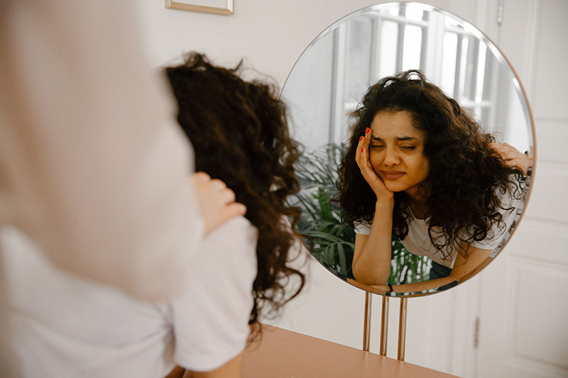 A young woman in front of a mirror expressing self-doubt and contemplation, while a hand rests on her shoulder.