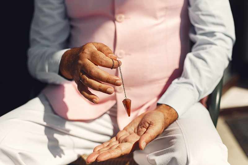 A man in soft pastel attire holding a gemstone pendulum over his palm.